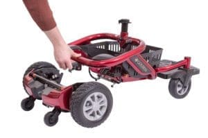 A person is assembling a Rental Power Wheelchair (Portable) with a red frame. The larger rear wheels and smaller front wheels are visible, as well as a rental basket at the back. A hand grips the scooters handle or component, preparing it for use.