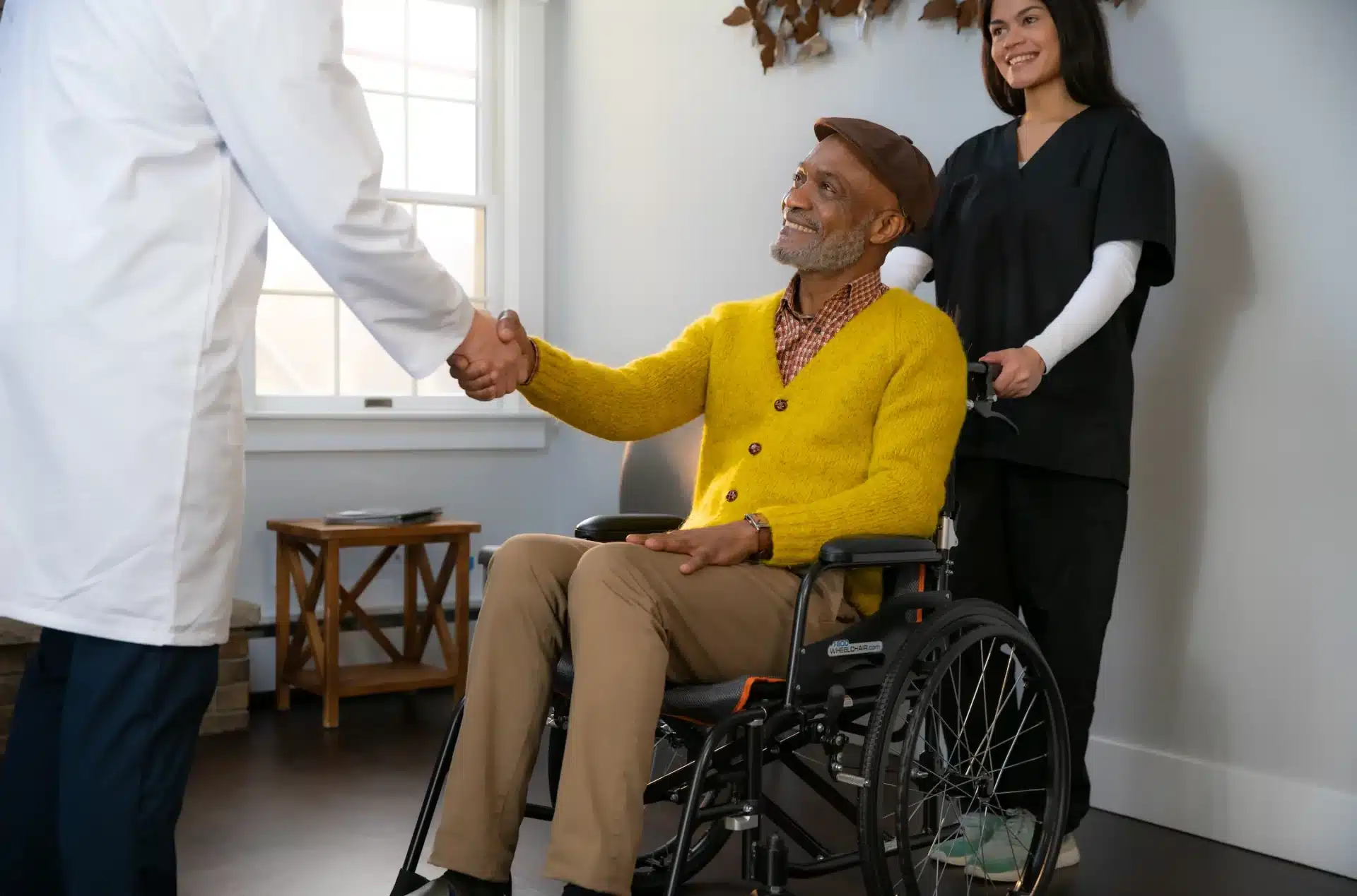 A man in a Feather Chair - 13.5 lbs, wearing a yellow sweater and brown pants, smiles as he shakes hands with a doctor. A nurse stands behind him, smiling, in a room filled with natural light from the window.