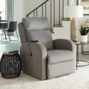 In a modern living room, the Golden Technologies PR763M EZ Sleeper Slim gray recliner is paired with a round black side table adorned with a textured lamp and small green plant. A staircase and dining area complete the inviting space.