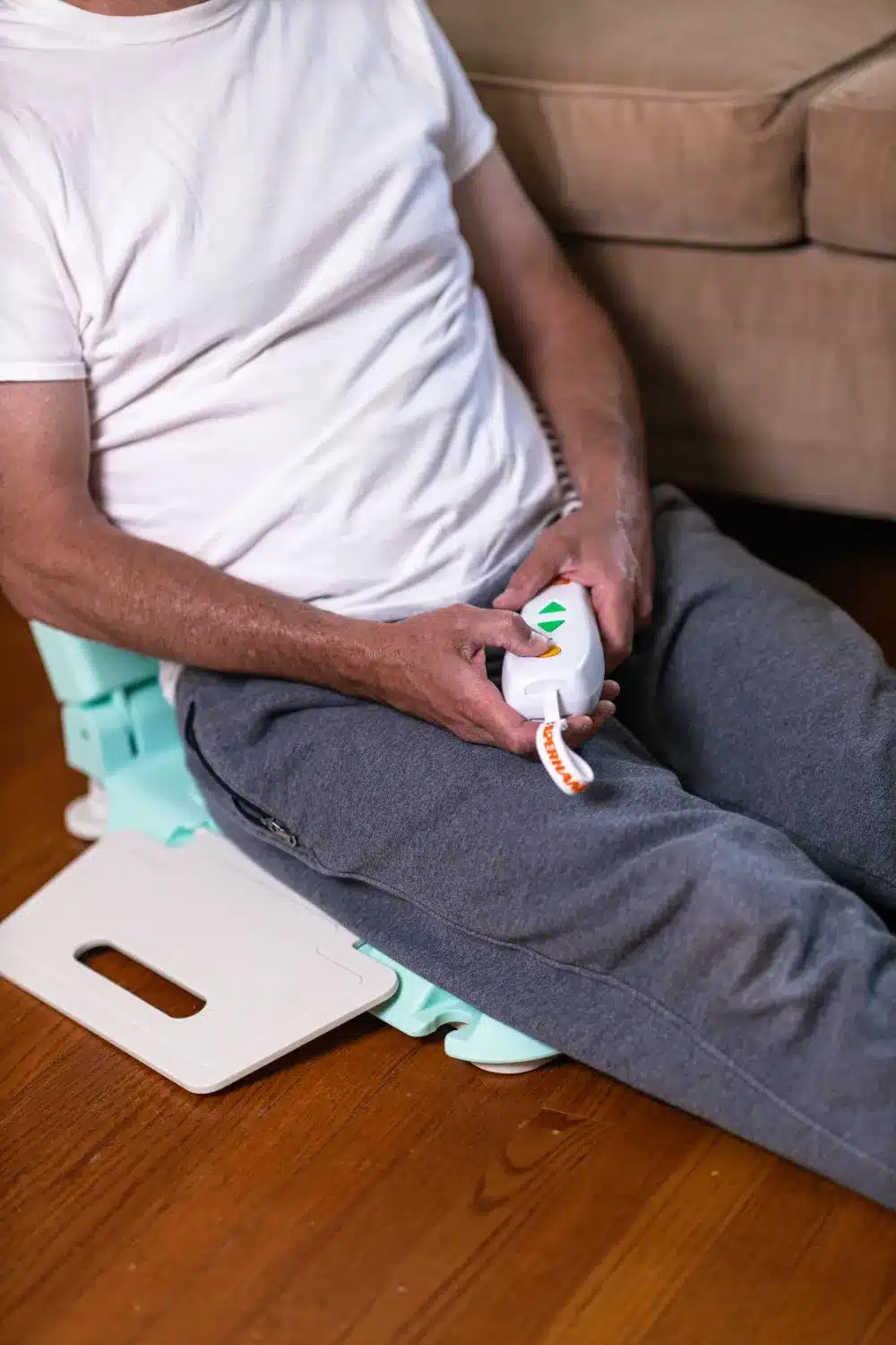 A person sits on the floor in a white t-shirt and gray pants, holding a remote control. Behind them, near the sofa, stands a SuperHandy GoRise LT Floor Bath Lift, subtly integrating assistive technology into their cozy living room with its wooden floors.