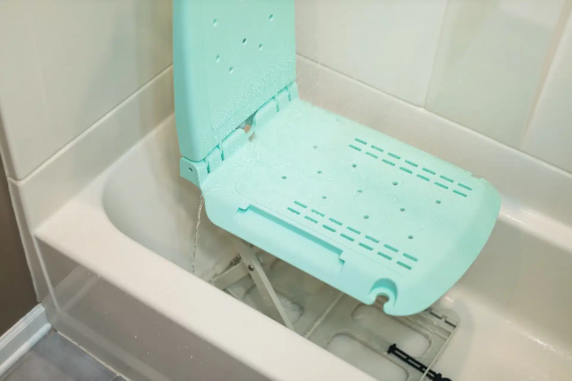 A light teal SuperHandy GoRise LT Floor Bath Lift with a backrest fits snugly in a white bathtub. Its foldable design has drainage holes and a sturdy metal frame. Water cascades over its surface, providing comfort and convenience for bathing.