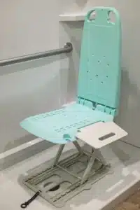 A teal folding shower seat is installed next to the SuperHandy GoRise LT Floor Bath Lift, featuring drainage perforations and a metal wall frame near a stainless steel grab bar. A visible side safety strap provides added security.