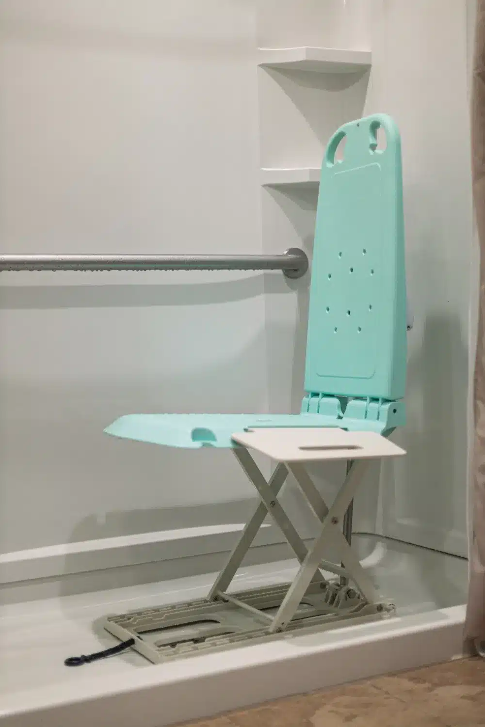 A light blue foldable shower chair, similar to the SuperHandy GoRise LT Floor Bath Lift, sits in a shower stall with white walls, built-in shelves, and a silver grab bar on the left. The chair features drainage holes and is supported by a white folding mechanism.
