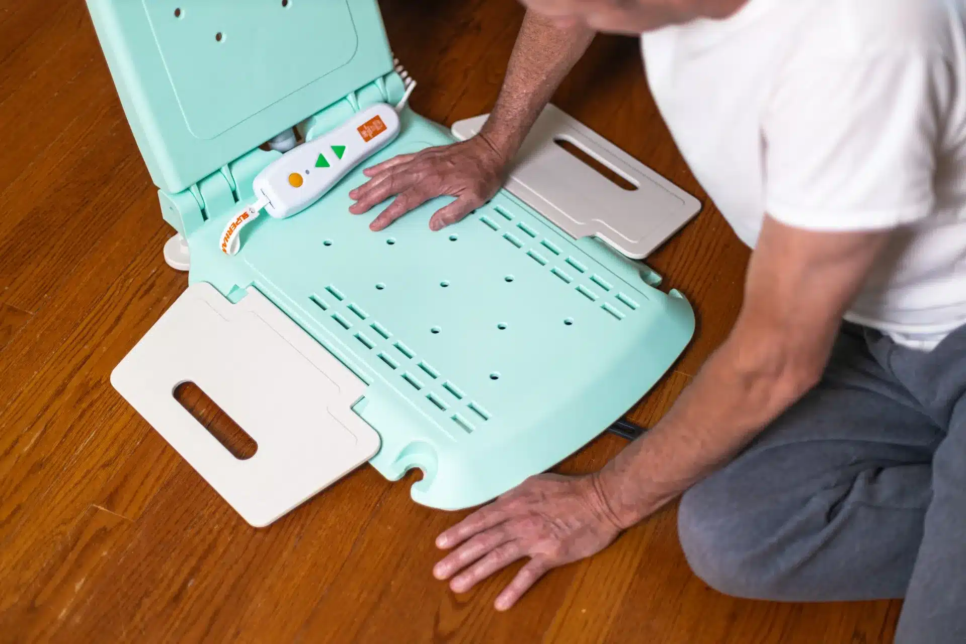 A person in a white shirt and gray pants assembles a SuperHandy GoRise LT Floor Bath Lift, adjusting its mint green and beige parts on the wooden floor. Their hands skillfully handle the bathroom aid bench, with tools scattered nearby.