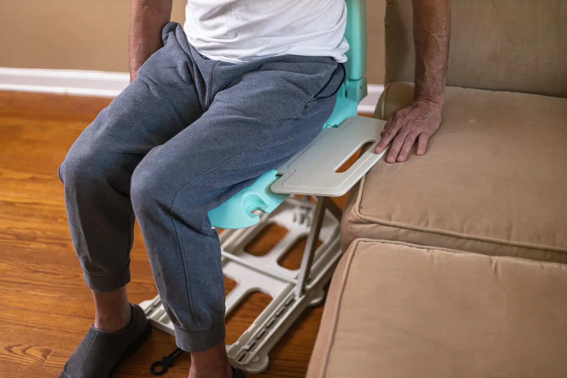 A person sits on a SuperHandy GoRise LT Floor Bath Lift beside a beige couch, dressed in a white shirt and gray sweatpants on a wooden floor, using the chairs armrest for support.