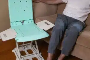 A person is adjusting a portable shower chair, akin to the SuperHandy GoRise LT Floor Bath Lift, while seated on a sofa. The chair has a light green seat and armrests. The individual wears a white shirt and gray pants comfortably.