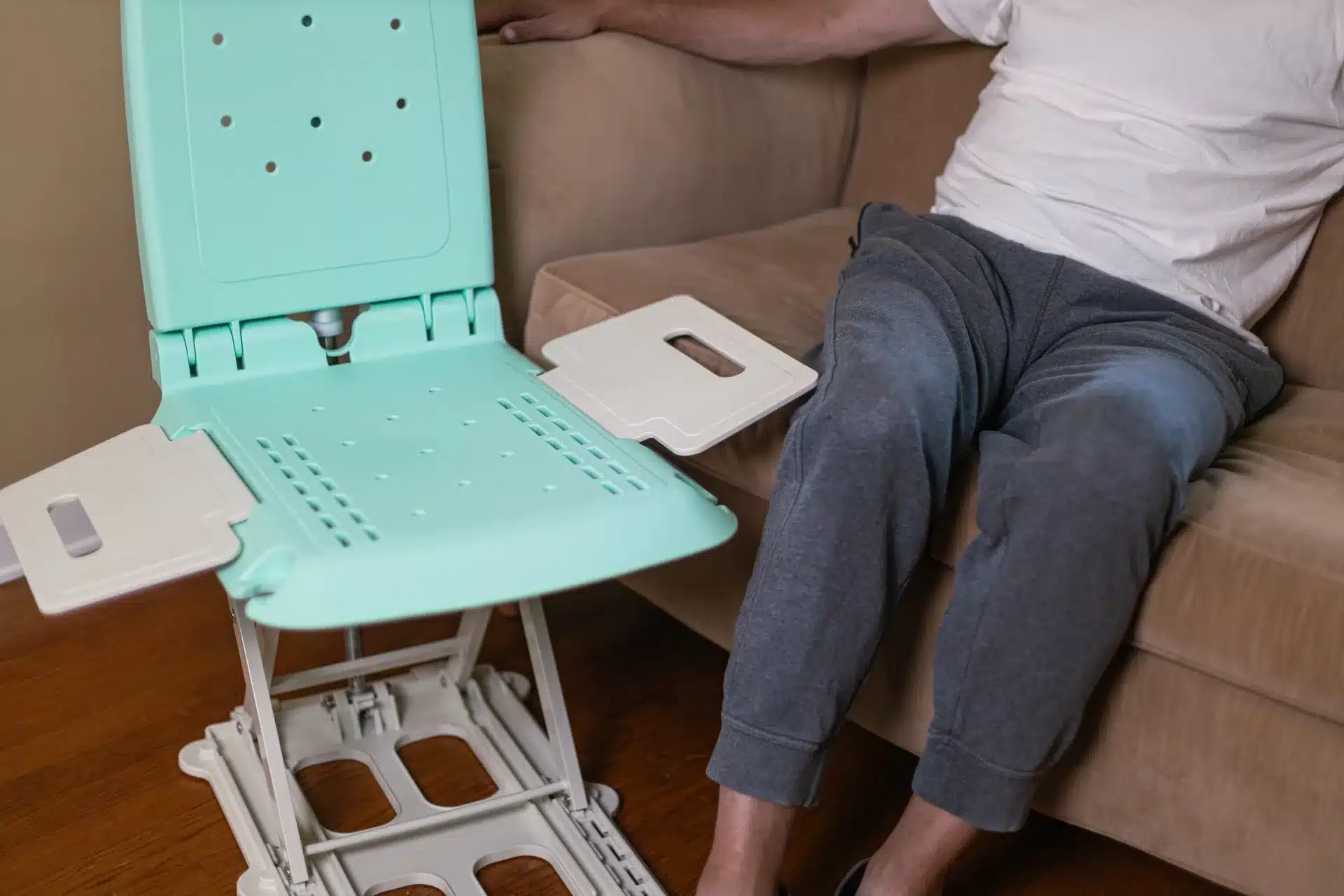 A person is adjusting a portable shower chair, akin to the SuperHandy GoRise LT Floor Bath Lift, while seated on a sofa. The chair has a light green seat and armrests. The individual wears a white shirt and gray pants comfortably.