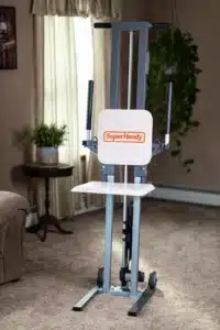 A SuperHandy GoRise FC Floor to Stand Mobility Lift enhances a cozy living room with its sleek design, complementing the brown furniture and side table. It features two vertical handles and a white platform, situated by a large window with curtains and flanked by a lush plant.