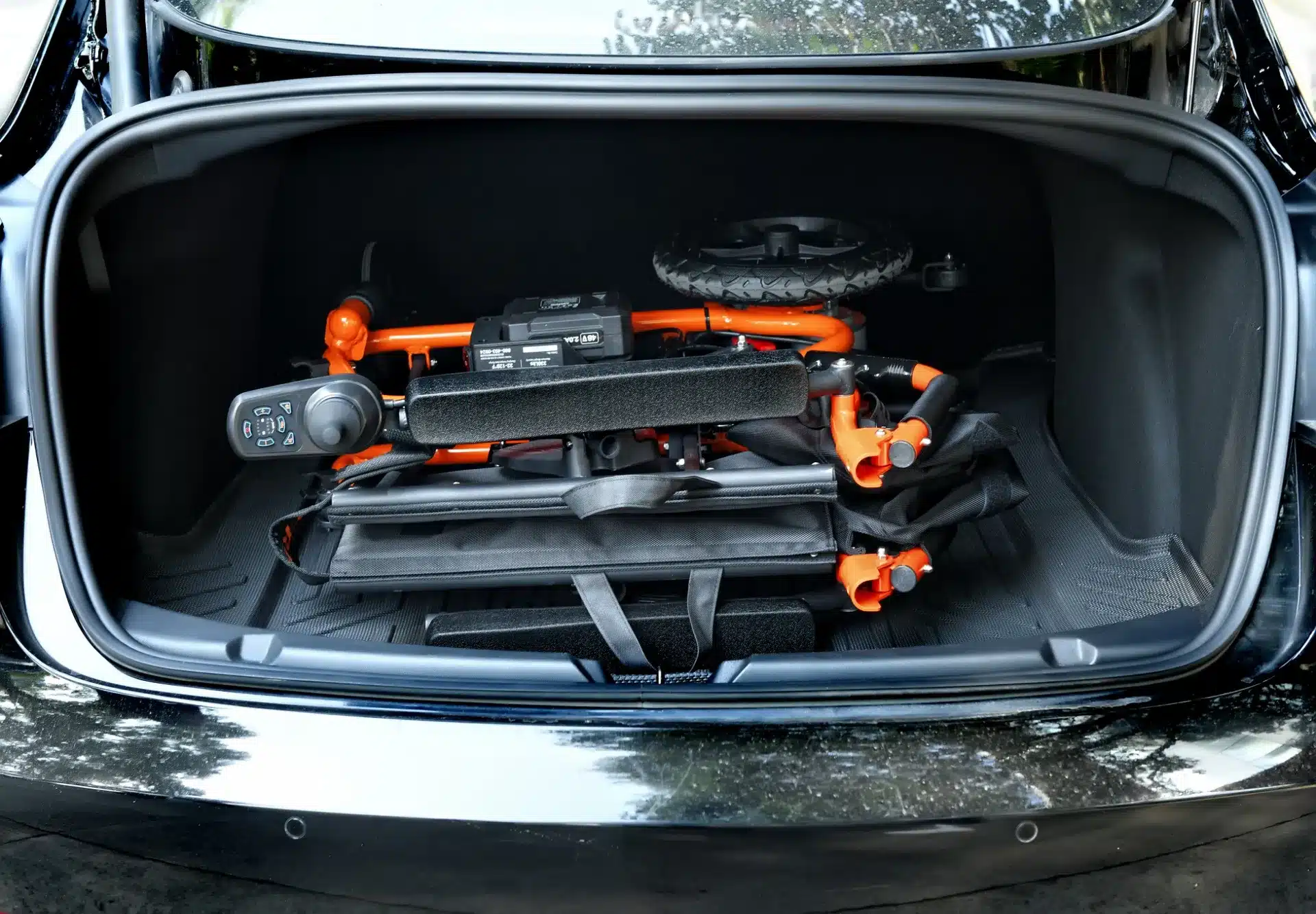 The SuperHandy Cares Portable Power Chair, featuring vibrant orange accents and intuitive controls, smoothly folds into a car trunk amid sunny outdoor scenery, highlighting the car’s roomy interior.