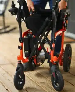 Someone is folding a vibrant orange SuperHandy Folding Electric Wheelchair on a wooden floor. The wheelchair, featuring visible wheels and a black seat, is being compactly collapsed for easy storage or transport.