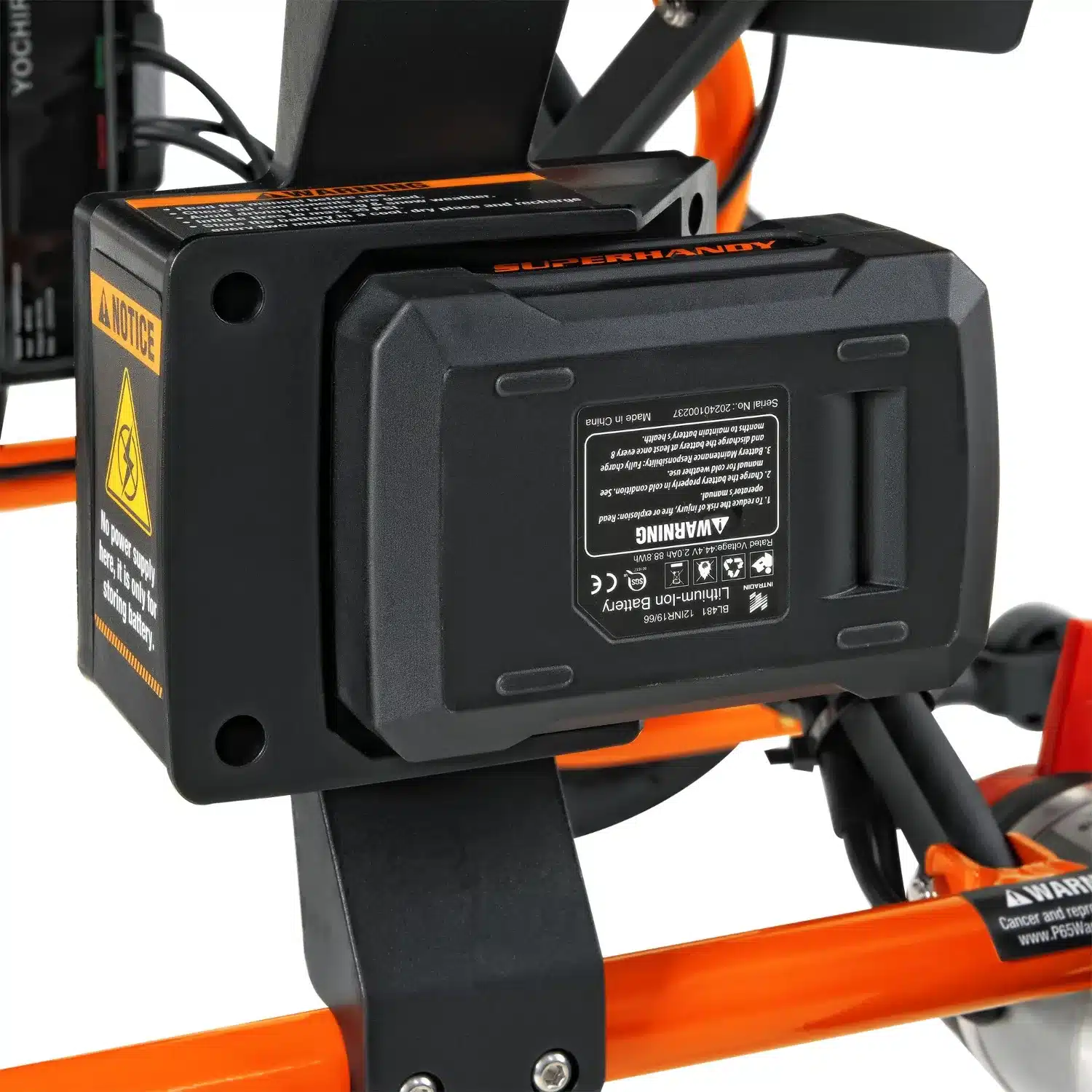 A black battery pack on the orange frame of the SuperHandy Cares Portable Power Chair shows safety labels and icons, emphasizing security. The chairs frame reveals connections and components of this innovative device.