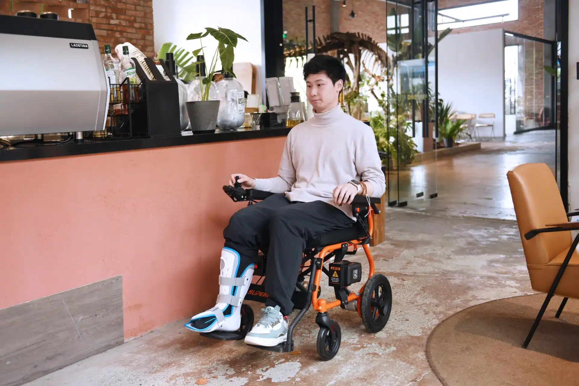 A person in a SuperHandy Cares Portable Power Chair smoothly maneuvers through a modern, spacious interior filled with lush greenery. They wear a light turtleneck paired with a medical brace on one leg.