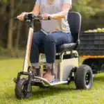 A person riding a sleek, seated electric scooter with SUPERMOTO emblazoned on the frame, towing a cart filled with apples across the grass. With SuperHandy Solutions enhancing mobility and independence, they wear jeans, a gray t-shirt, and brown boots. Trees stand serenely in the background.