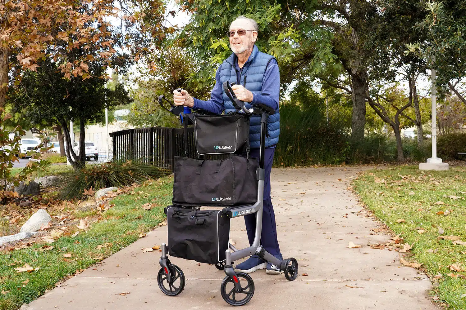 An elderly man in a blue vest and jeans navigates the park path with ease using his UPWalker® EZ Lite Tool-free Folding Backsaver Rollator, enjoying the sunny autumn trees and crisp weather.