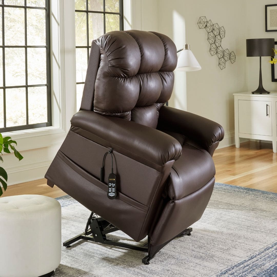 In a well-lit room with large windows, the Golden Cloud PR 517 recliner in rich brown leather is elevated by its lift mechanism. A remote control hangs at the side, and it is complemented by a gray rug and a white ottoman nearby.