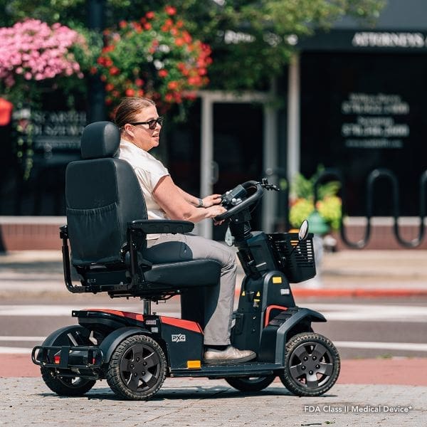 A person wearing sunglasses and a ponytail speeds through city streets on a sleek black and red PX4 All Terrain Outdoor Mobility Scooter, with greenery and blurred storefronts enhancing the scenes dynamic energy.