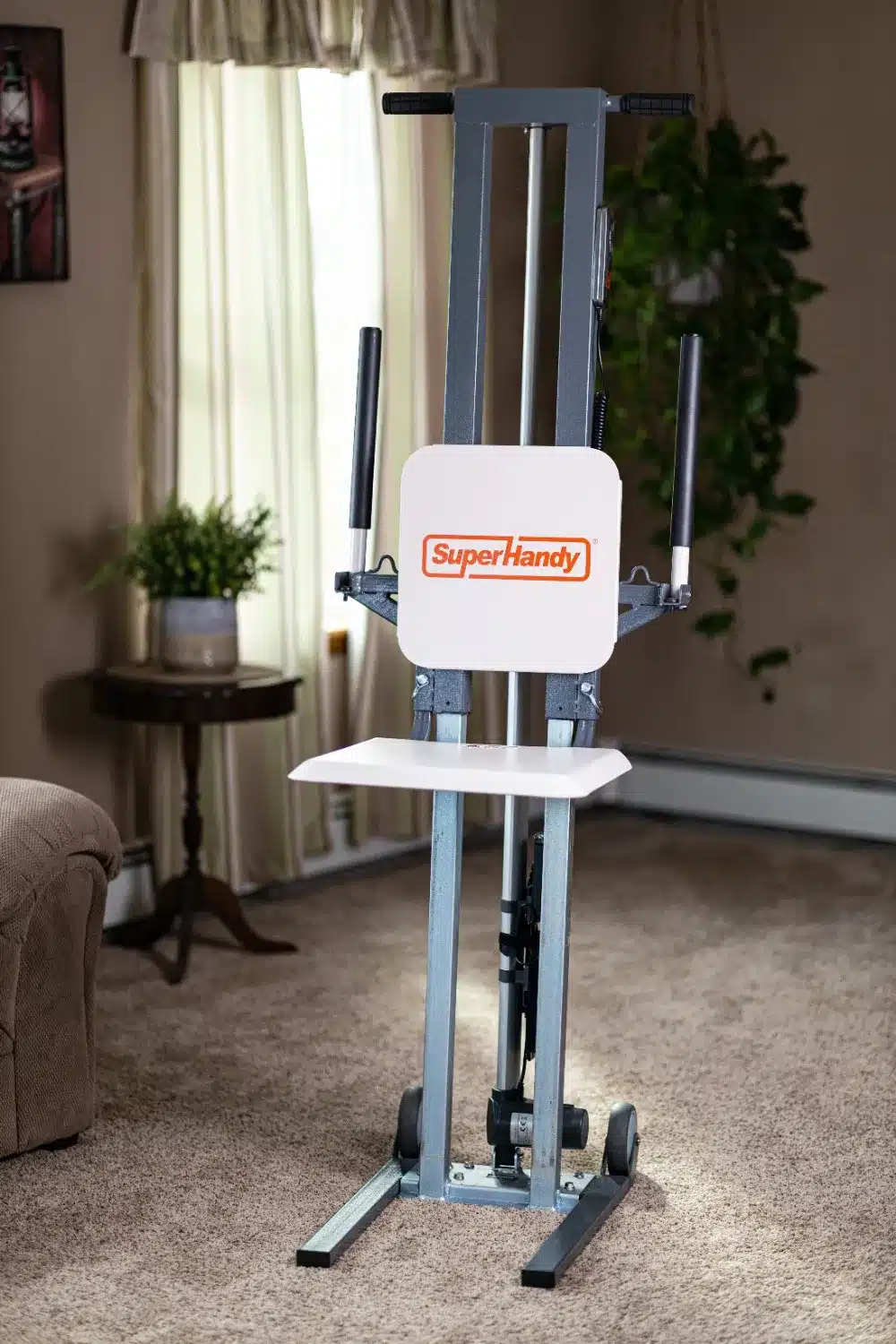 A SuperHandy GoRise FC Floor to Stand Mobility Lift enhances a cozy living room with its sleek design, complementing the brown furniture and side table. It features two vertical handles and a white platform, situated by a large window with curtains and flanked by a lush plant.