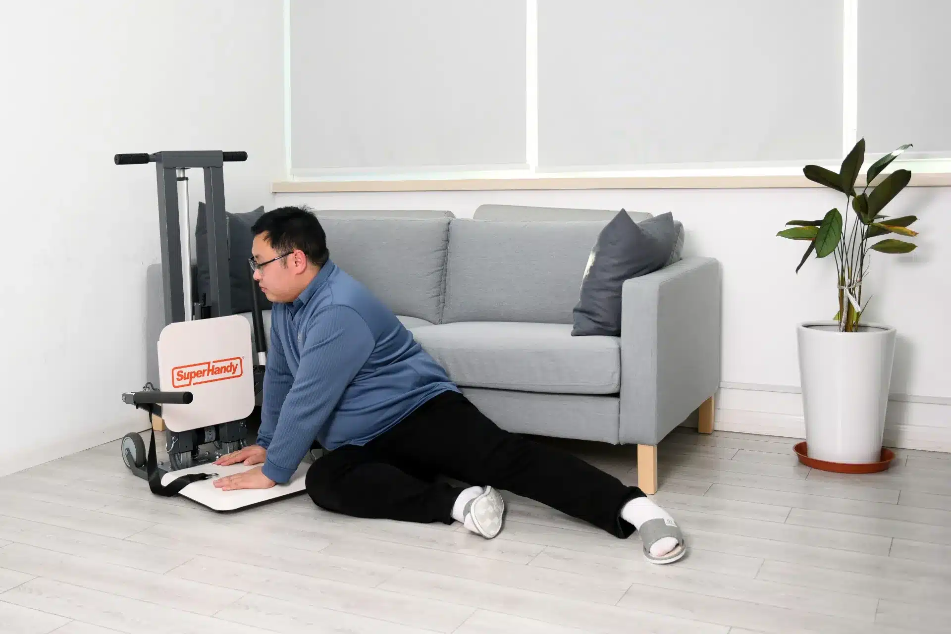 A person in a blue jacket and black pants sits on the floor next to a SuperHandy GoRise FC Floor to Stand Mobility Lift in a cozy living room, near a grey sofa with cushions. A potted plant adds natural decoration nearby.