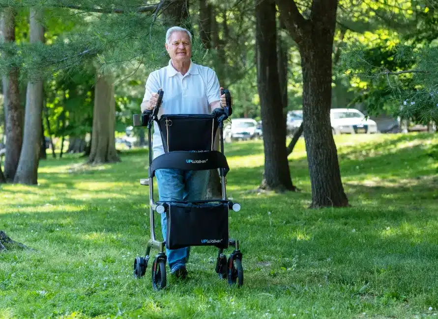 An older adult in a white shirt and jeans relishes a sunny day in the park with their UPWalker Premium Lite, amid lush greenery and trees, with parked cars visible behind them.