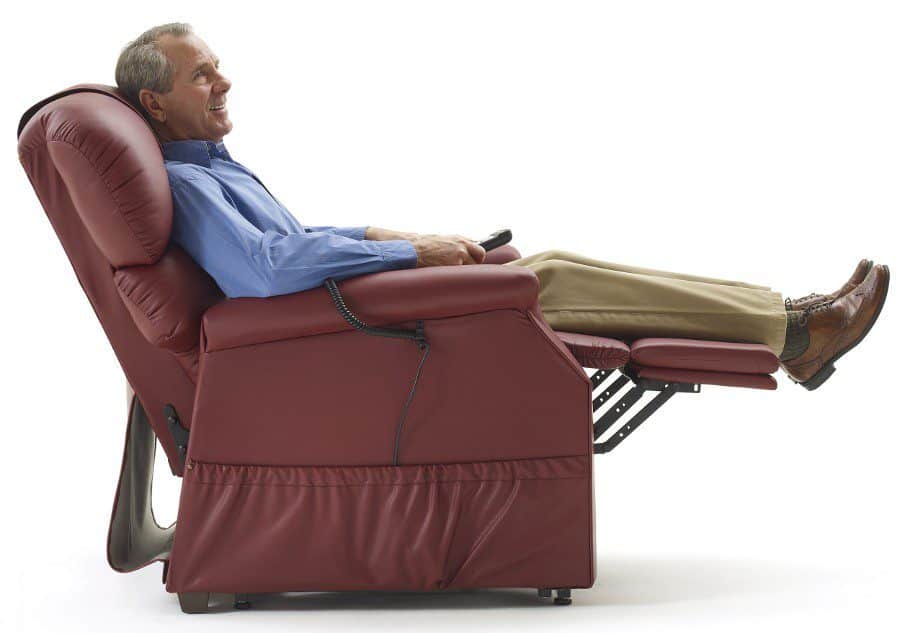 A man in a blue shirt and beige pants is sitting comfortably in a red reclining chair, holding a remote control. The chair is fully reclined, with his feet elevated, and he appears relaxed against a plain white background.